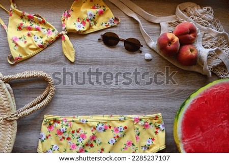 Floral yellow bikini, wicker bag, retro sunglasses, seashell, bag with peaches and cut watermelon on wooden background. Top view, copy space. 