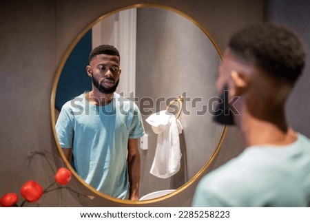 Unhappy upset 30s African American man looking in mirror, standing in bathroom at home, sad depressed black guy feeling dissatisfied with his appearance. Midlife crisis in men Royalty-Free Stock Photo #2285258223