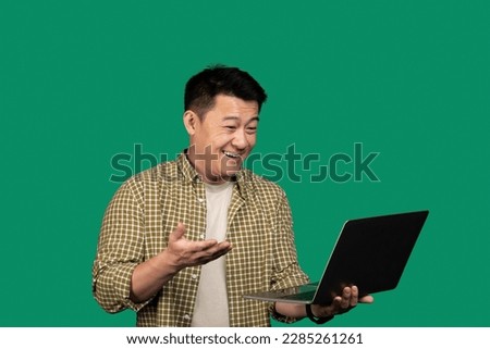 Positive asian middle aged man having video call with friend or family via laptop, standing over green background, looking at computer screen, smiling and gesturing, copy space