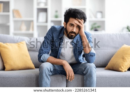 Portrait Of Depressed Young Indian Man Sitting On Couch At Home, Pensive Upset Eastern Guy Touching Head And Looking Away, Suffering Depression, Life Problems Or Mental Breakdown, Copy Space Royalty-Free Stock Photo #2285261341