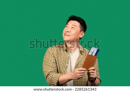 Portrait of asian middle aged man holding passport with plane boarding pass tickets, looking aside at copy space on green studio background. Overseas tourism, abroad vacation concept Royalty-Free Stock Photo #2285261343