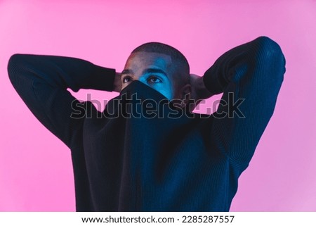 Cool and fashionable man. Young Black guy hiding his face in his sweatshirt and holding hands behind his neck. Studio pink background. High quality photo