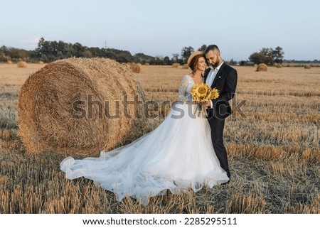 Wedding portrait of the bride and groom standing against the background of a dry field, hugging and looking into the camera lens. Red-haired bride with open shoulders. Stylish groom. Summer