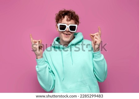 young curly guy in mint hoodie and sunglasses dances and snaps his fingers on pink isolated background, man in blue sweatshirt moves his hands to the music at the party