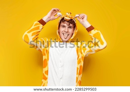 young joyful guy in funny baby giraffe pajamas shows tongue and holds ears on yellow background, man in animal cosplay clothes, pajama party concept
