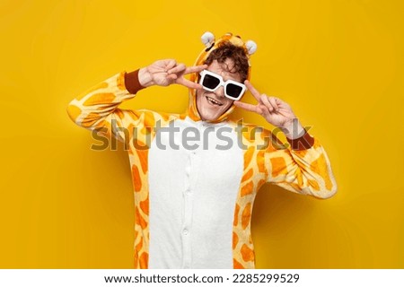 young joyful guy in funny baby giraffe pajamas and glasses is dancing on yellow background, man in animal cosplay clothes shows peace gesture, pajama party concept