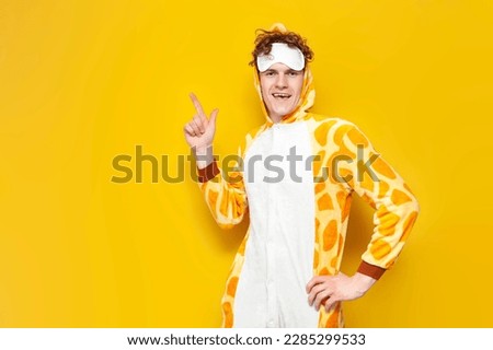 young joyful guy in funny baby giraffe pajamas and sleep mask shows his hand to the side on yellow background, man in animal cosplay clothes advertises copy space