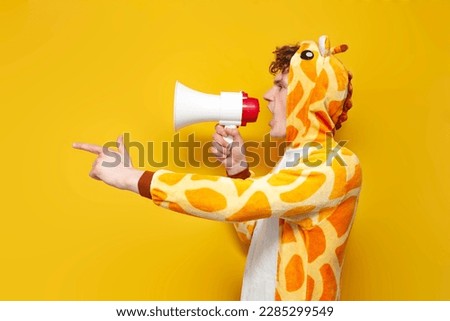 young joyful guy in funny children's giraffe pajamas speaks into megaphone and points his hand to the side on yellow background, man in animal cosplay clothes announces and shouts