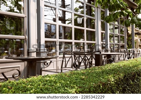Empty outdoor cafe - metal forged furniture fenced by green hedge. Tables and chairs at restaurant terrace