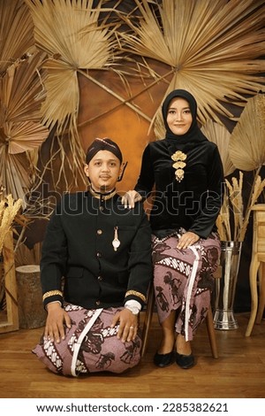 A couple wearing Javanese traditional clothes, wearing a black shirt and dress. male model sitting on the floor and female sitting on a chair. Very elegant Javanese traditional photo. natural photo ba