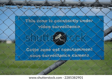 Airport Environment - Airport fence with a warning sign in English and French language "This zone is constantly under video surveillance" in front of blurred background 