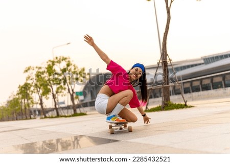 Cool Asian girl skating on longboard skate at park on summer holiday vacation. Stylish young woman having fun urban outdoor lifestyle practicing extreme sports skateboarding on city street at sunset.