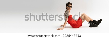 full length of slim tattooed woman in black boots and red corset dress sitting on grey background, banner