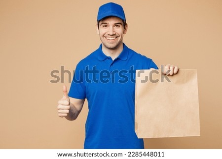 Delivery guy employee man wear blue cap t-shirt uniform workwear work as dealer courier hold brown clear blank craft paper takeaway bag mock up show thumb up isolated on plain light beige background