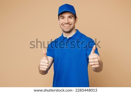 Professional satisfied delivery guy employee man wear blue cap t-shirt uniform workwear work as dealer courier showing thumb up like gesture isolated on plain light beige background. Service concept Royalty-Free Stock Photo #2285448013