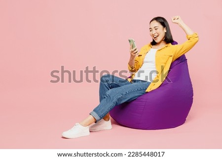 Full body young woman of Asian ethnicity wears yellow shirt white t-shirt sit in bag chair hold in hand use mobile cell phone do winner gesture isolated on plain pastel light pink background studio Royalty-Free Stock Photo #2285448017