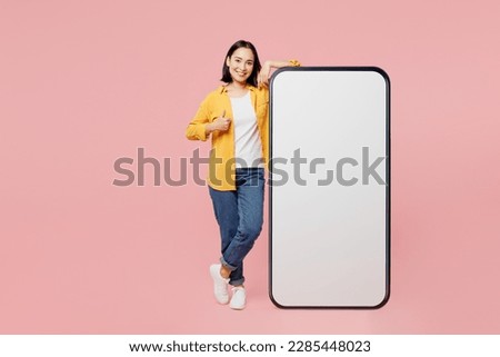 Full body fun young woman of Asian ethnicity wear yellow shirt white t-shirt big huge blank screen mobile cell phone smartphone with area show thumb up isolated on plain pastel light pink background