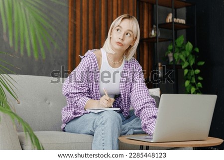 Young IT woman in casual clothes work hold use laptop pc computer write in notebook look aside sit on grey sofa stay at home hotel flat rest relax spend free time in living room People lounge concept