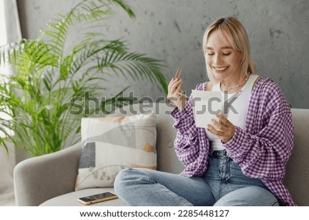 Young fun woman wear casual clothes eat Chinese food cuisine hold takeaway carton container box sits on grey sofa couch stay at home hotel flat rest relax spend free spare time in living room indoor