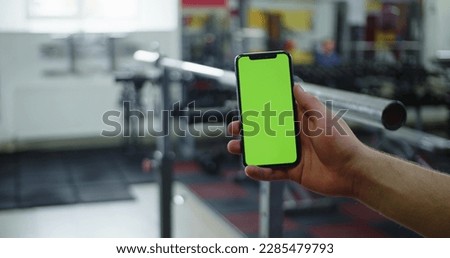Male hand holding smartphone in gym, fitness app for bodybuilding, green screen