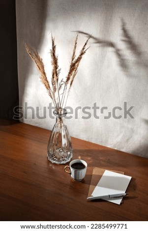 Aesthetic summer interior still life with cup of coffee, paper cards and dry grass in glass vase on a wooden table, floral sunlight shadows on a wall, morning business concept