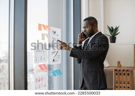 Focused multiracial businessman in smart suit receiving phone call while standing near glass panel with graphs and charts in corner office. Senior executive listening to worker's speech over mobile.