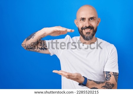 Hispanic man with tattoos standing over blue background gesturing with hands showing big and large size sign, measure symbol. smiling looking at the camera. measuring concept. 