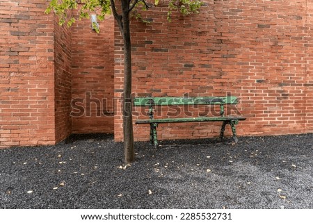An old green bench stands next to a green tree in front of a brick wall on a stone chipping background Royalty-Free Stock Photo #2285532731