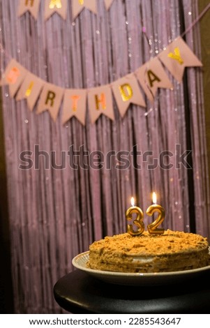 Cake with candles with numbers 3 and 2 on a background of pink rain with flags Happy Birthday in gold.
