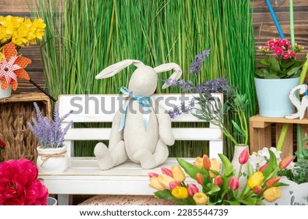 Easter bunny is sitting on white bench against background of green grass , festive spring decor in photo studio. Greetings and postcards. Spring photo shoot. Festive decor. Design of photo zones.