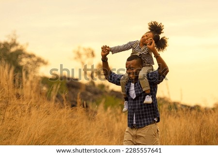 Happy African family child daughter riding the neck father and running on meadow nature on silhouette lights sunset.  Travel and Family Concept