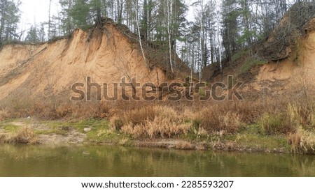 River landscape in late autumn. The high river bank with sandy bluffs is overgrown with birches and pines. Tall dry grass stands by the water. The bank is reflected in the water. Overcast weather Royalty-Free Stock Photo #2285593207