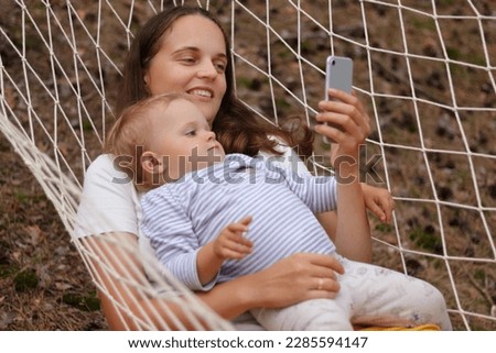 Portrait of Caucasian woman with her infant kid in forest, mother and daughter lying in hammock, female using mobile phone, watching cartoons together with her child.