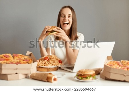 Portrait of overjoyed woman laptop sitting at table with computer and fast food, rejoicing ending diet, eating junk food, isolated over gray background.