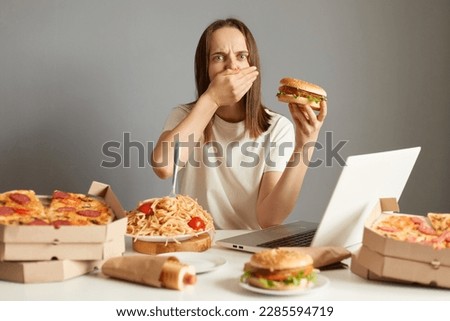 Portrait of sick overeating woman sitting at table in front of notebook isolated over gray background, holding sandwich, feels bad and nausea, covering mouth with palm.