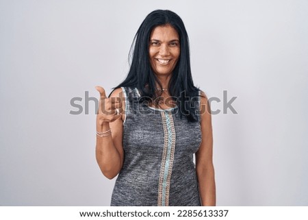 Mature hispanic woman standing over white background doing happy thumbs up gesture with hand. approving expression looking at the camera showing success. 