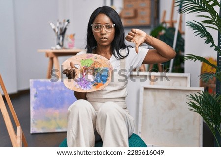 African young woman holding painter palette with angry face, negative sign showing dislike with thumbs down, rejection concept 