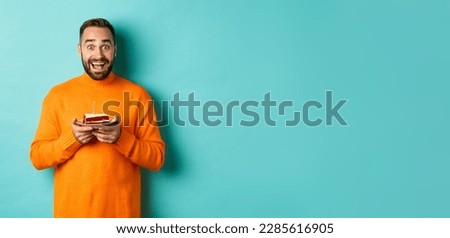 Happy adult man celebrating birthday, holding bday cake with candle and smiling, standing against turquoise background.