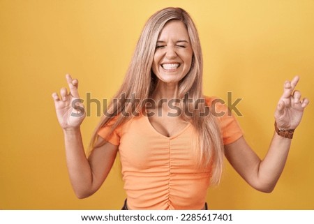 Young woman standing over yellow background gesturing finger crossed smiling with hope and eyes closed. luck and superstitious concept. 
