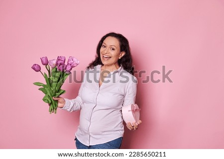 Happy smiling pregnant woman smiles, looking at camera, posing with a cute present in a heart shaped gift box and a bouquet of purple tulips for Mother's or International Women's Day, isolated on pink