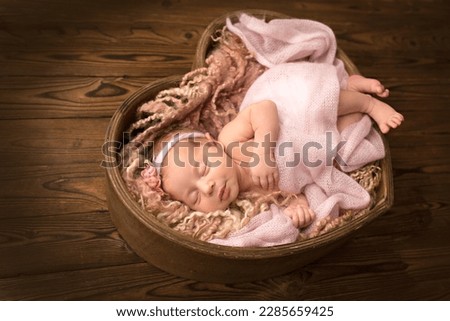 Newborn baby girl sleeping in a pink wrap with a white bandage with a pink flower on her head. Wooden basket in the shape of a heart on a wooden brown background. Little girl 7 days, one week.