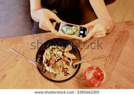Top view of unrecognizable woman sitting at wooden table near black plate with vegetable salad and glass with red cocktail, fork, knife in restaurant cafe, holding smartphone, taking pictures of food.