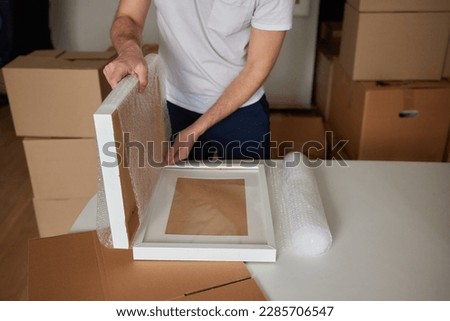 A young man crouched down looking through a moving box, filled with clothes, books and a picture frame