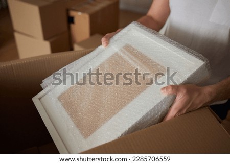 A young man crouched down looking through a moving box, filled with clothes, books and a picture frame