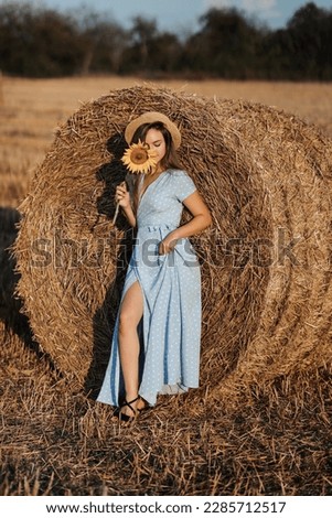 Portrait of a young girl. A girl in a blue dress and a hat holds a sunflower flower covering her face against a background of hay bales. Long straight hair. Nice color. Summer