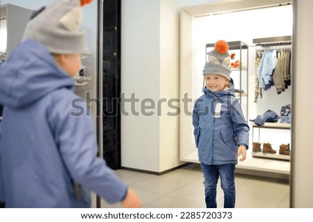 Cute little boy is trying on a new coat and hat in front of a mirror in a store or shopping mall. Fashionable warm kids clothing for autumn or winter. The child likes to go shopping with parents