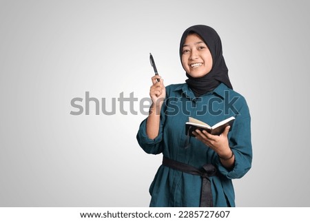 Portrait of excited Asian muslim woman with hijab writing on note book and pointing up with pen. Advertising concept. Isolated image on white background