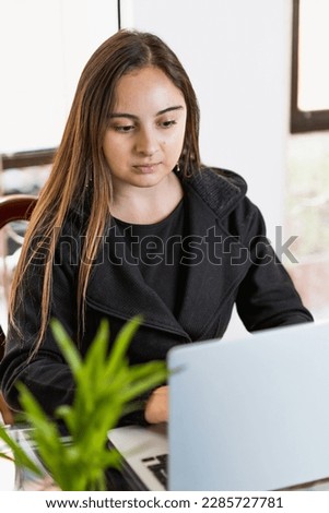 Young latin woman focused on work in front of laptop near windows, vertical photo