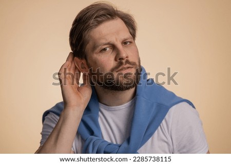 I cant hear you. What. Man trying hear you, looking confused and frowning, keeping arm near ear for louder voice, asking to repeat to hear information, deafness. Young guy isolated on beige background Royalty-Free Stock Photo #2285738115