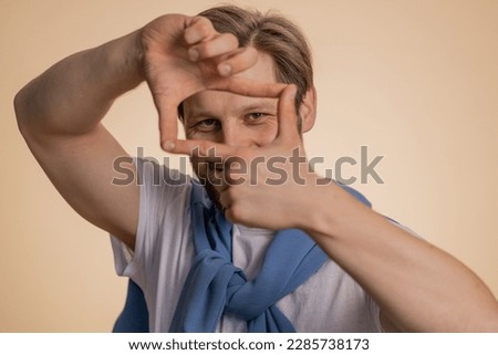 Man photographer gesturing picture frame with hands, looks through fingers and focusing on interesting moment, imitating zoom and cropping nice image. Young guy isolated on beige studio background